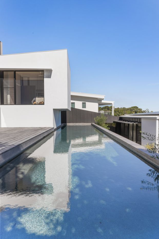 Exterior architectural photograph of a contemporary multi-level residence with crisp white rendered walls, dark privacy screens and extensive glazing. A long narrow reflecting or swimming pool runs between the buildings, with timber decking along the left edge and a clear blue sky above.