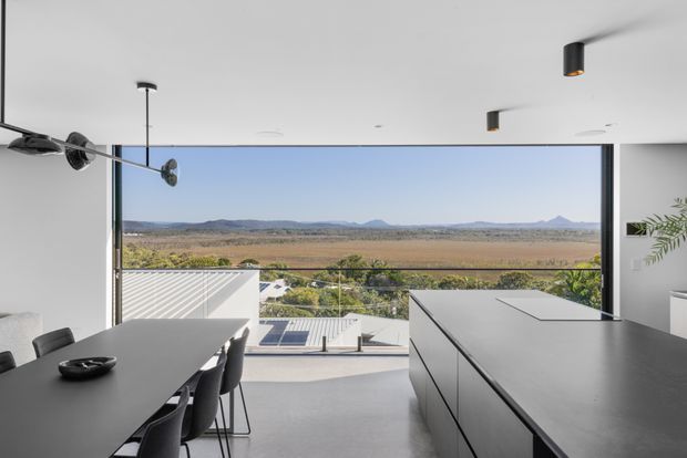 Contemporary open-plan interior with a dark dining table and chairs beside a large minimalist kitchen island. Full-height glazing opens to an expansive rural landscape and distant hills, while a covered outdoor terrace and pool area are visible beyond. The space has crisp white walls and ceiling, dark cabinetry and furnishings, polished concrete flooring, and restrained black architectural lighting.