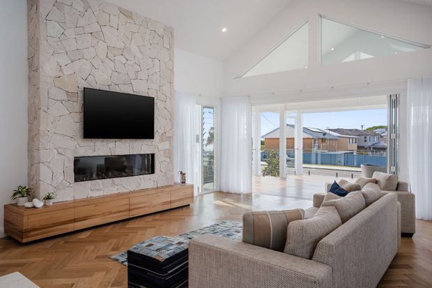Bright contemporary living room with a large stone-clad fireplace wall, wall-mounted television, built-in timber media cabinetry, herringbone timber flooring, and a large neutral sectional sofa. The room opens to a balcony or outdoor view through expansive glazing, with a high angled ceiling and clerestory-style windows.