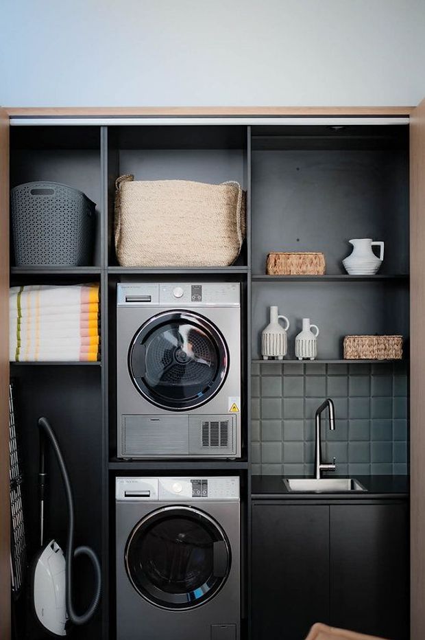 Compact, dark-toned laundry room with stacked front-loading washer and dryer integrated into black open shelving and cabinetry. The room includes a deep grey-green square-tile splashback, black sink cabinet with a tall black tap, timber-framed joinery, folded towels, woven baskets, ceramic vessels, and a vacuum cleaner.