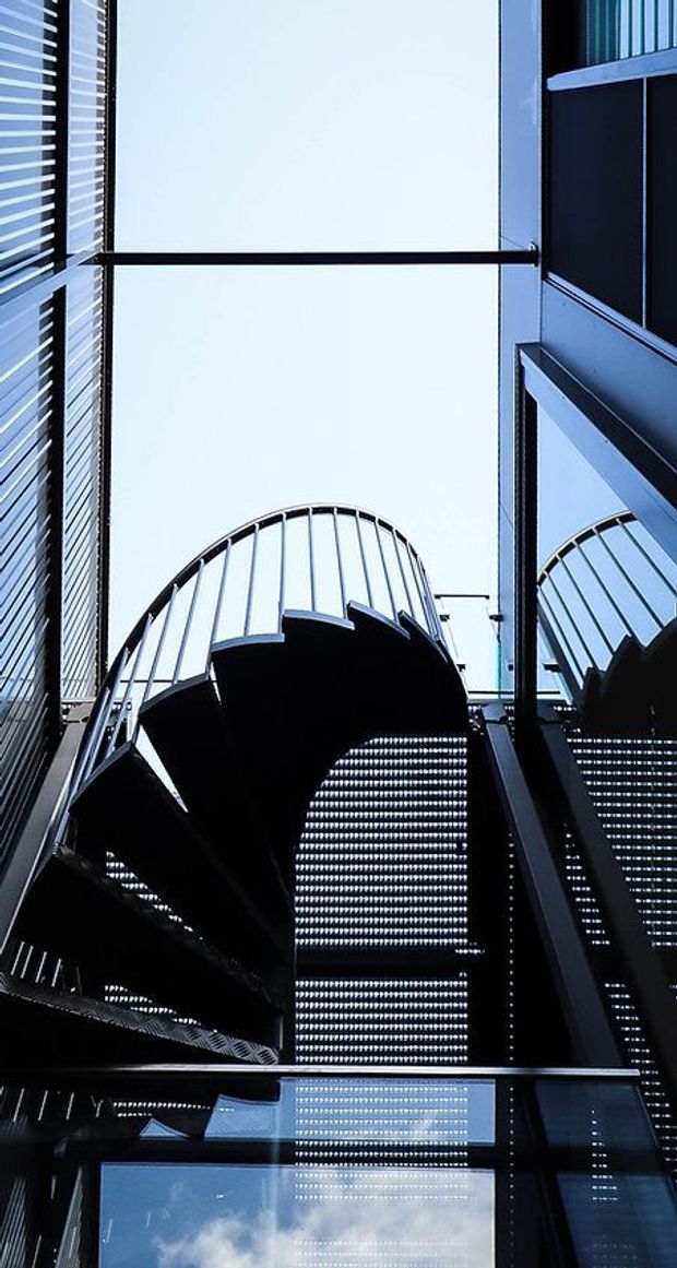 Upward-looking view within a dramatic modern stairwell or atrium, featuring a sculptural curved metal staircase, perforated metal stair treads, dark steel framing, reflective glazing, and strong geometric lines against a bright sky.