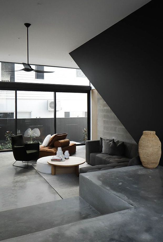 A moody contemporary living room with polished grey concrete floors and a dramatic black angular feature wall or stair volume. The room has large black-framed glazed doors and windows, a dark grey sofa, black lounge chair, brown leather armchair, round timber coffee table, sculptural pendant and ceiling fan, with a raised concrete platform and woven floor vase.