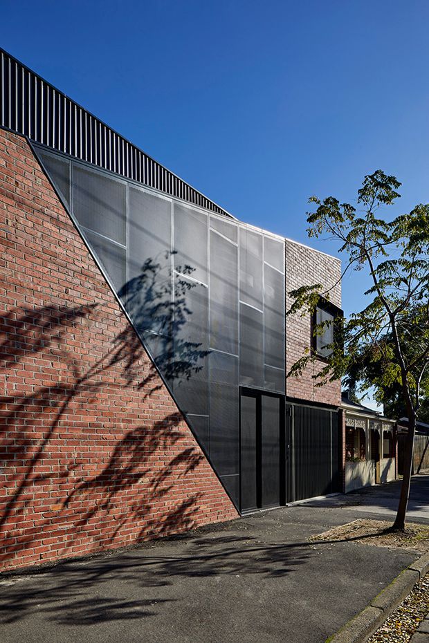 Street-facing view of a contemporary urban building facade combining exposed red brickwork, dark metal screening and framing, and translucent corrugated or perforated panels. The angular facade rises from a sloping brick wall to a higher screened roof edge, with a narrow black entrance and garage-like opening at ground level. Mature street trees, pavement, and a clear blue sky surround the building.