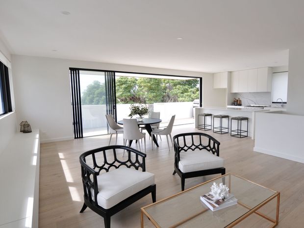 Bright, open-plan contemporary living and dining area with a compact white kitchen at the rear. The room features pale timber flooring, white walls and cabinetry, black-framed full-height sliding glazing opening to a balcony or terrace, a small dining setting, black-and-white accent lounge chairs, and a glass coffee table with a brass-toned frame.