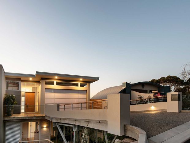 Exterior architectural photograph at dusk showing a contemporary multi-level residence on a sloping urban site. The house has a flat projecting roof, rendered walls, extensive glazing, a timber-toned entry door, a large sectional garage door, upper-level terrace screening, and a curved dark roof form on the adjoining structure. A driveway and retaining walls occupy the foreground.