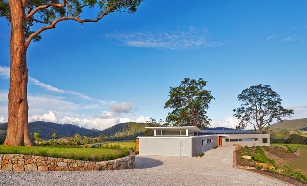 A low, modern single-storey residence set in a rural landscape, viewed from a broad gravel driveway. The home has a minimalist horizontal composition with pale rendered or masonry walls, dark charcoal roof planes, a large garage door, clerestory-style upper glazing, and a sheltered entry. Stone retaining walls, landscaped garden beds, mature trees, rolling hills, and a bright blue sky surround the property.