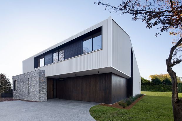 A contemporary two-storey residence with a strong geometric form, white vertically ribbed cladding, dark timber-look garage and entry doors, and a contrasting grey stone-clad lower volume. A long band of dark-framed upper-level glazing sits beneath a projecting box-like roof form. The house is set within a landscaped suburban lawn with a paved driveway and mature trees.
