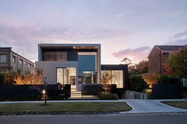 Street-facing dusk photograph of a contemporary multi-level residence with a bold rectangular flat-roofed form, dark charcoal and light rendered facade, extensive floor-to-ceiling glazing, and a lower single-storey wing. The home sits behind a landscaped front garden with dark fencing, a paved driveway, and mature neighbouring houses visible on either side.