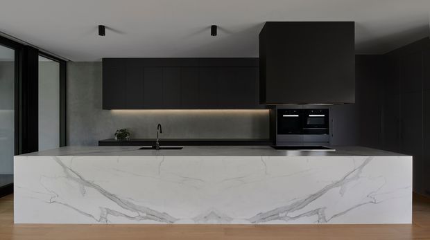 A dark, minimalist contemporary kitchen viewed from the living side, dominated by a large white veined stone island with an integrated sink and black tap. Full-height dark cabinetry runs along the rear and side walls, with integrated black appliances beneath a large box-like rangehood. Black-framed glazing is visible to the left, while warm timber flooring adds contrast.