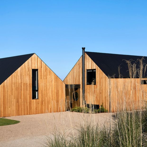 Exterior view of a contemporary single-storey residence composed of two steeply gabled volumes. The facades are clad in warm vertical timber boards, with narrow dark-framed windows and black pitched roofs. A gravel driveway and native ornamental grasses frame the building under a clear blue sky.