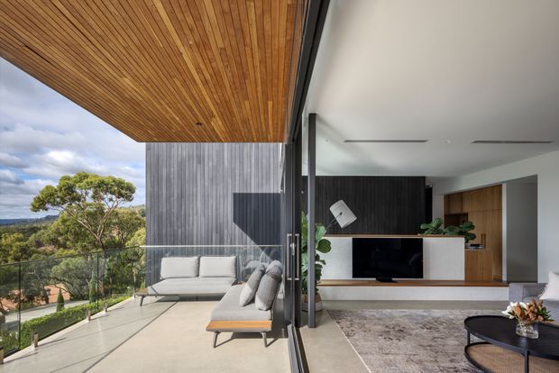 A contemporary open-plan living area opening onto a spacious elevated terrace. The interior features a dark vertical timber feature wall, white built-in media cabinetry, warm timber flooring and joinery, and large glass sliding doors. Outside, the terrace has a timber-lined soffit, concrete paving, glass balustrade, and outdoor seating overlooking a leafy suburban landscape.