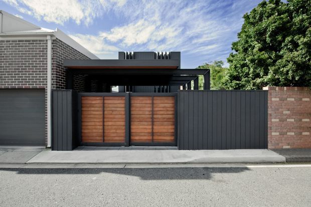 Street-level view of a contemporary residential frontage featuring a dark charcoal slatted garage and gate structure, warm timber garage doors, exposed brick walls, and a covered entry or carport-like volume. The facade combines horizontal and vertical lines with a flat roof form and strong contrasting materials.