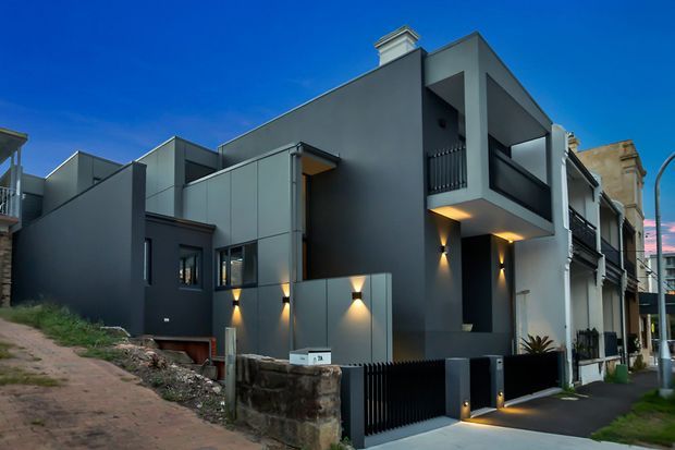 Contemporary multi-level urban residence with a dark grey rendered facade, projecting upper-storey balcony, recessed entry, vertical metal balustrades, wall-mounted exterior lighting, and a narrow driveway. The home sits within a dense suburban streetscape alongside older neighbouring buildings.