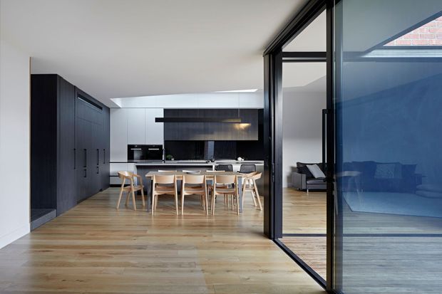 A contemporary open-plan kitchen and dining area viewed through a large black-framed glazed opening. The room features a long island with pale stone or light-coloured benchtop, dark full-height cabinetry and a dark splashback, a timber dining table with light timber chairs, and wide light timber flooring. An adjoining living area is visible through the glazing, with strong indoor-outdoor visual connection and abundant natural light.