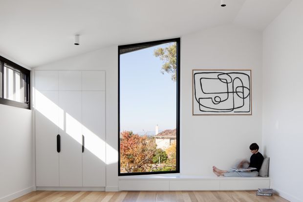 Bright minimalist interior room with white built-in cabinetry, a large black-framed vertical window, light timber flooring, and a built-in window seat. A person is seated reading beside a simple abstract line artwork. Strong natural light creates a calm, airy atmosphere.