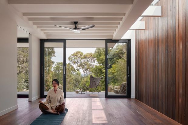 A spacious, serene interior with polished dark timber flooring, a dramatic full-height vertical timber feature wall, extensive black-framed glazing opening toward a leafy landscape, and a white exposed-beam ceiling with an integrated ceiling fan. A person sits on a yoga mat in the foreground, while a lounge chair is visible beyond the glazing.