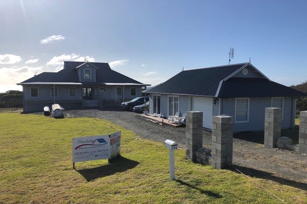 Exterior view of a low-rise rural residential property with two dark-roofed, grey-clad buildings, gravel driveway, unfinished block fence pillars, and a broad grassy foreground under a clear sky. The main house has gabled roof forms and dormer windows, while the secondary building appears to be a garage or ancillary dwelling.