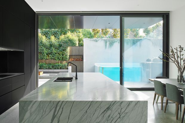 Contemporary open-plan kitchen with a large veined stone island and dark full-height cabinetry. Floor-to-ceiling sliding glass doors open the space toward a bright outdoor pool courtyard, while a dining table and chairs sit to the right. The room has a clean, luxurious, indoor-outdoor character with strong natural light.
