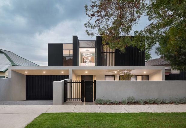 Street-facing exterior view of a contemporary two-storey residence with a flat roof, crisp white rendered walls, dark vertical screening and cladding, an integrated garage, and a recessed entry behind a slatted gate. Large upper-level glazing and warm exterior lighting provide contrast against the dark facade elements, while a low front boundary wall and landscaped verge frame the property.