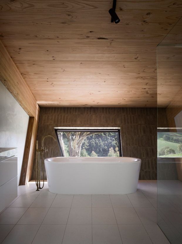 A contemporary bathroom featuring a freestanding white bathtub beneath a warm timber-lined sloped ceiling. Large angled glazing overlooks a green landscape, while dark textured wall tiles, pale floor tiles, a brass floor-mounted bath tap, and a full-height mirror create a refined, nature-connected interior.