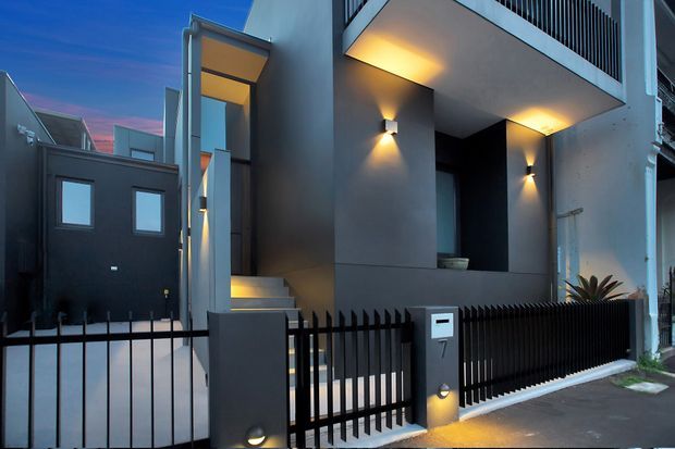 Contemporary multi-level urban townhouse facade at dusk, featuring smooth dark and light rendered walls, recessed balconies and entry canopy, warm wall-mounted lighting, a black metal picket fence, front gate, and a compact driveway or entry path. The image focuses on the building exterior and street-facing entry.