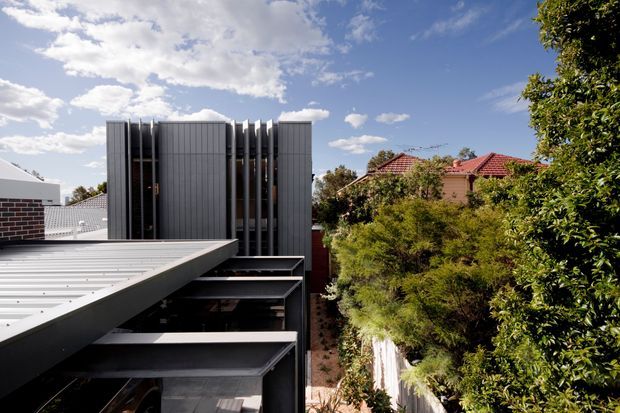 Exterior architectural photograph of a contemporary multi-level building viewed from above or across a roof terrace. The building has a dark grey metal-clad facade with strong vertical fins and projecting horizontal slabs, set among established suburban vegetation and neighbouring red-tiled houses under a partly cloudy sky.
