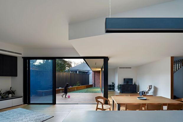 Contemporary open-plan living and dining area with a timber dining table and chairs, a media unit, and expansive black-framed sliding glass doors opening onto a timber deck and enclosed courtyard. The room features a white palette, pale flooring, a suspended dark linear pendant, integrated cabinetry, and a visible stairway at right.
