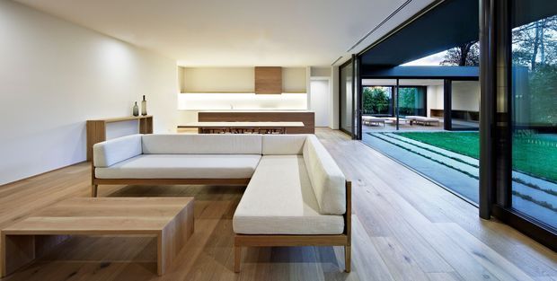 A refined contemporary living room with a large light upholstered sectional sofa, low timber coffee tables, timber flooring, and minimalist built-in shelving and fireplace joinery. Full-height black-framed glazing opens the room toward a landscaped courtyard and covered outdoor area, creating a strong indoor–outdoor connection. The palette is warm and restrained, with white walls, pale timber, and dark window frames.