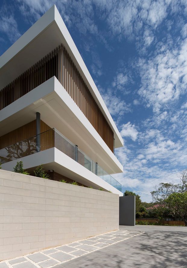 Low-angle exterior view of a multi-storey contemporary residence with strong white horizontal slab projections, expansive balconies with clear glass balustrades, warm timber vertical screening, and a pale masonry boundary wall. The image emphasizes the sharp geometric roofline and facade against a bright blue sky, with a paved driveway and landscaped planting at ground level.