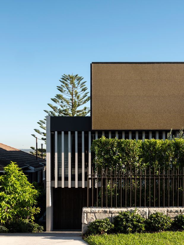 Street-facing view of a contemporary multi-level residential facade with a dark rendered lower volume, slender vertical fins, a large upper facade clad in tightly spaced warm-toned horizontal slats, and a black metal fence set above a pale stone retaining wall. Dense hedging and established conifer trees frame the property, with neighbouring rooftops visible at the edges.