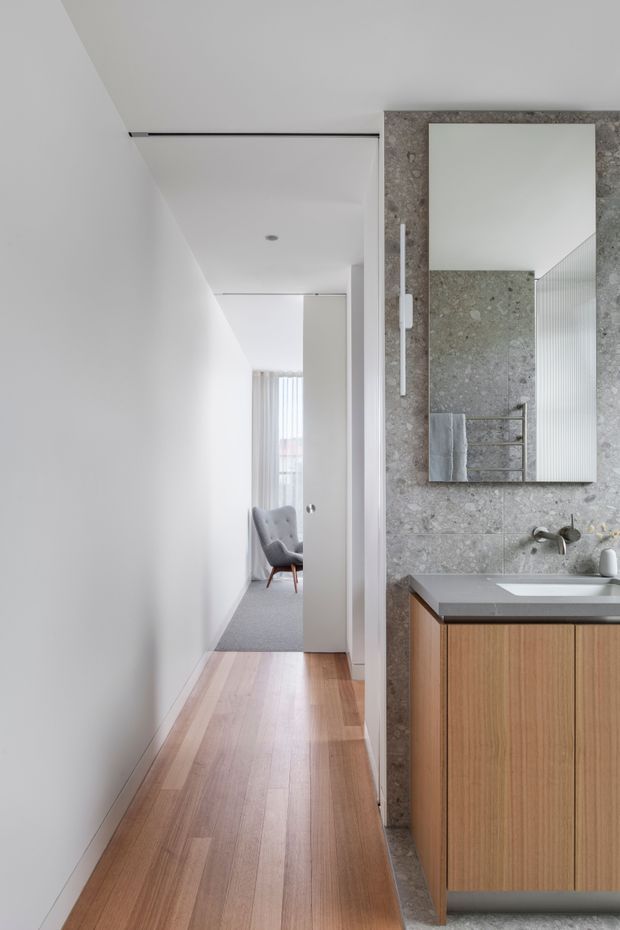 A bright, narrow contemporary hallway opening directly onto a compact bathroom vanity area. The corridor has smooth white walls and ceiling, warm timber flooring, and a grey stone-tiled bathroom wall with a large frameless mirror, wall-mounted tapware, grey benchtop, and vertical timber vanity cabinetry. A softly furnished room is visible at the end of the hall.
