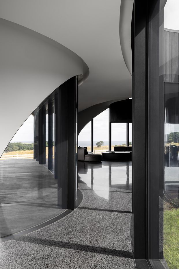 A dramatic contemporary interior with sweeping curved white ceilings, expansive dark-framed glazing, and a polished dark aggregate floor. The open-plan space contains a low dark seating area and round table, with views toward a coastal or rural landscape. Strong reflections, sculptural arches, and black vertical window mullions create a refined, minimalist atmosphere.