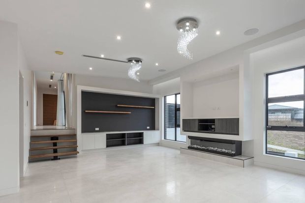 Bright contemporary open-plan living room with polished light flooring, white walls and ceiling, a dark built-in media wall, and a sculptural built-in fireplace. A short set of timber-edged stairs leads toward a hallway, while large black-framed windows and recessed lighting create an airy, refined interior.