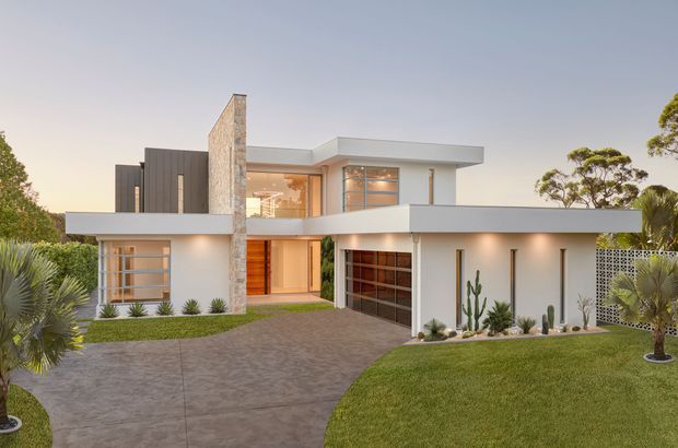 Exterior view of a contemporary two-storey residence with strong horizontal roof and balcony forms, white rendered walls, dark vertical cladding, a tall natural stone chimney or feature wall, extensive glazing, and an integrated double garage. The home is approached by a broad paved driveway and surrounded by manicured lawn and drought-tolerant landscaping.