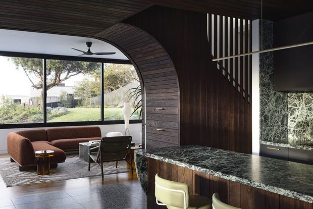 A sophisticated open-plan living and bar area with a curved timber-lined ceiling and arched timber partition framing expansive glazing. The room combines dark timber wall panelling, a dramatic veined stone bar countertop, charcoal tiled flooring, and mid-century-inspired lounge furniture. A sculptural pendant hangs above the bar, while a staircase with vertical balustrade slats is visible behind it. The glazing opens views to a landscaped garden.