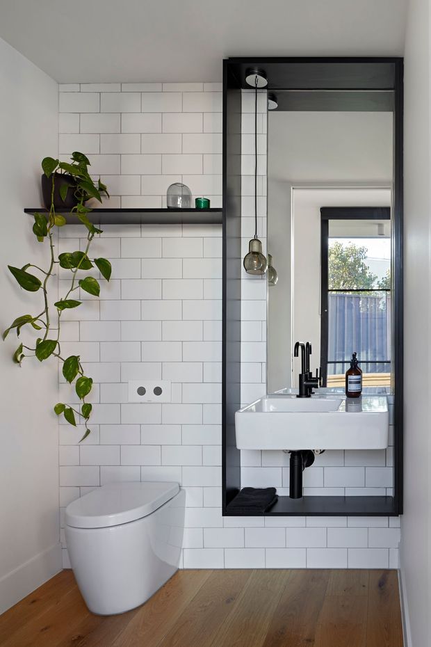 Compact contemporary bathroom with white subway-tile walls, a wall-mounted white basin, black tapware, a black-framed full-height mirror, wall shelf, pendant lights, and a modern white toilet. A trailing indoor plant adds greenery, while warm timber flooring contrasts with the monochrome fixtures.