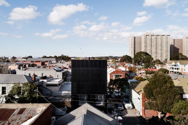 Elevated view across a dense suburban/urban neighbourhood, showing low-rise houses and commercial or industrial rooftops in the foreground with a prominent tall apartment building in the background. The scene is photographed outdoors in bright daylight under a partly cloudy sky.