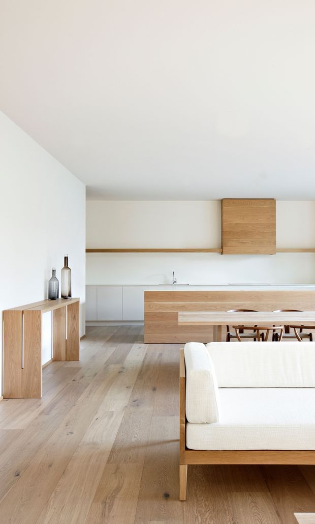 Bright open-plan interior combining a minimalist kitchen, dining area and living space. The room features white walls and ceiling, pale wide-plank timber flooring, light timber cabinetry and joinery, a simple dining table with timber chairs, and a white upholstered sofa in the foreground. A slim timber shelf runs along the rear wall, with understated decorative vessels on a low timber console.