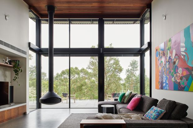 Contemporary living room with a large double-height glazed wall opening onto greenery, a dark sectional sofa, colourful artwork, built-in media joinery, and a sculptural suspended fireplace. The room has a warm timber-lined ceiling, dark structural window framing, and a bright indoor-outdoor connection.
