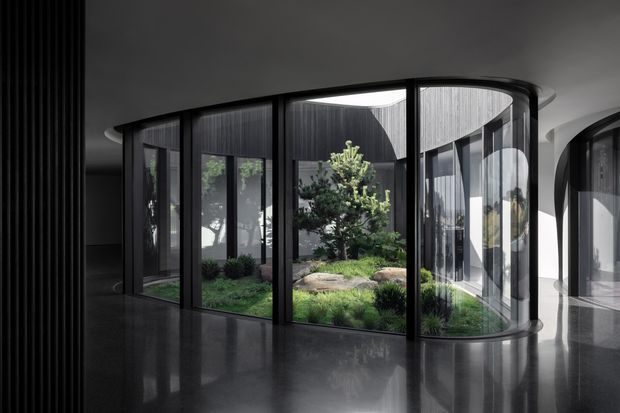A dramatic contemporary interior featuring a curved, floor-to-ceiling glazed enclosure wrapped around a landscaped internal courtyard. Black slender window frames, dark ribbed wall and ceiling surfaces, and a highly reflective dark floor create a moody, gallery-like atmosphere, while the planted courtyard introduces natural greenery and daylight. The image also shows a secondary curved glazed opening at right.