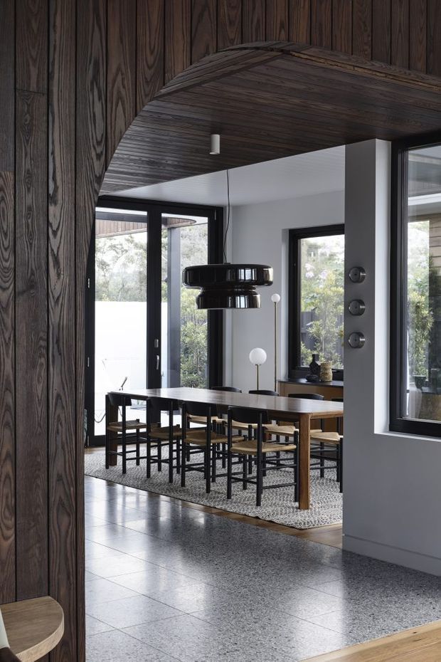 A refined dining area viewed through a dark timber-lined opening. The room features a long rectangular timber dining table with black-framed chairs, a large black pendant light, floor-to-ceiling glazing, dark window frames, and a light grey wall palette. The ceiling and curved opening are clad in richly grained dark timber, while the floor is finished in grey terrazzo. The overall composition is contemporary with strong mid-century-inspired furniture and lighting.