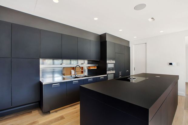 Contemporary kitchen with extensive matte dark cabinetry, integrated appliances, a stainless-steel splashback and sink area, and a large dark island with a smooth benchtop. Light timber flooring and white walls and ceiling provide contrast, with recessed downlights and minimal detailing.