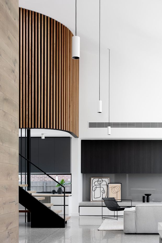 A tall, refined contemporary living area with a double-height ceiling and a dramatic curved vertical timber-slat feature wall. The space includes a black sculptural staircase with glass balustrade, minimalist suspended white pendant lights, dark built-in cabinetry, a low white media console, abstract artwork, and pale upholstered seating. A kitchen or dining area is visible beyond the staircase. The palette is predominantly white, black, grey, and warm timber, with polished flooring and strong vertical architectural detailing.