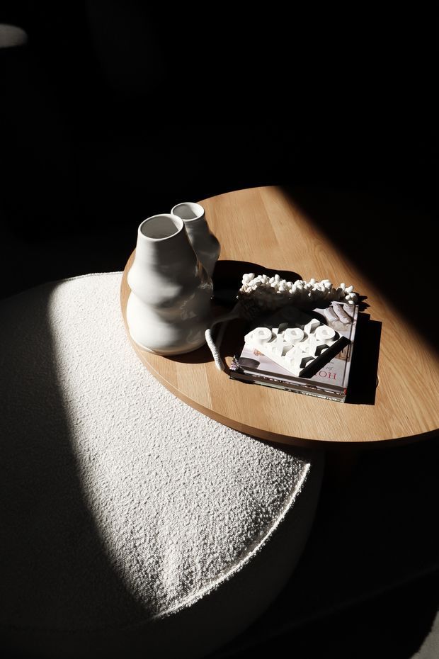 A moody, close-up interior styling scene featuring a light timber oval coffee table over a plush off-white textured rug. The table holds sculptural white ceramic vessels, a pale textured decorative object, and stacked books. Strong directional sunlight creates dramatic shadows against a dark surrounding interior.
