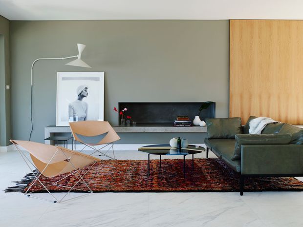 A refined mid-century-inspired living room with a low built-in media and display unit, olive-green wall, warm timber cabinetry, dark green leather sofa, sculptural lounge chairs, black metal coffee table, and a richly patterned rug over a pale stone floor. A large framed portrait and minimalist floor lamp complete the calm, curated interior.