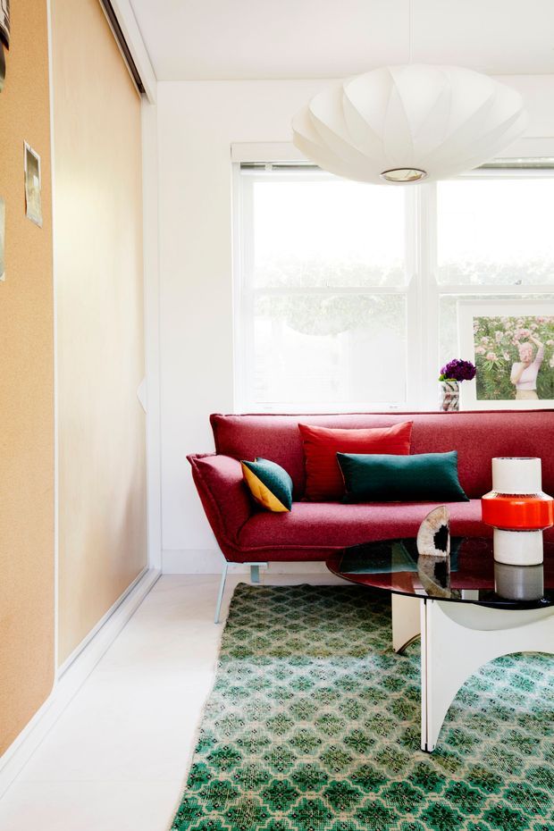 Bright, compact living room with a vivid red upholstered sofa, jewel-toned cushions, a patterned green rug, a black glass coffee table, and a sculptural white pendant light. White walls, pale flooring, large windows, and a warm tan feature wall create a colourful mid-century-inspired interior.