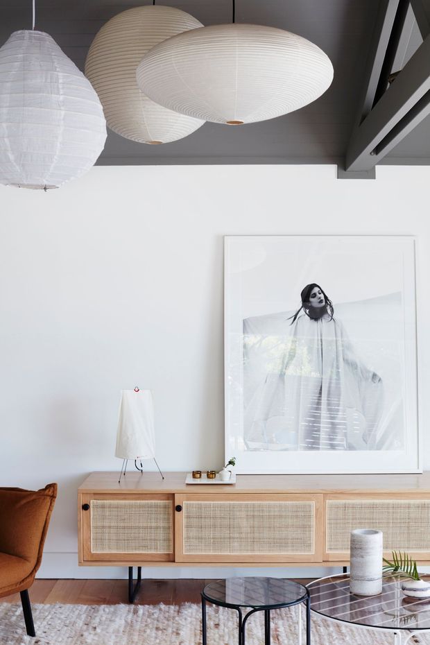 A bright, refined living room with white walls and a dark exposed-beam ceiling. Three oversized cream paper pendant lights hang above a low timber sideboard with woven cane-front cabinetry. A large monochromatic framed artwork leans above the sideboard, while a caramel lounge chair, glass-and-black-metal coffee tables, neutral rug, ceramics, and indoor plants complete the space.