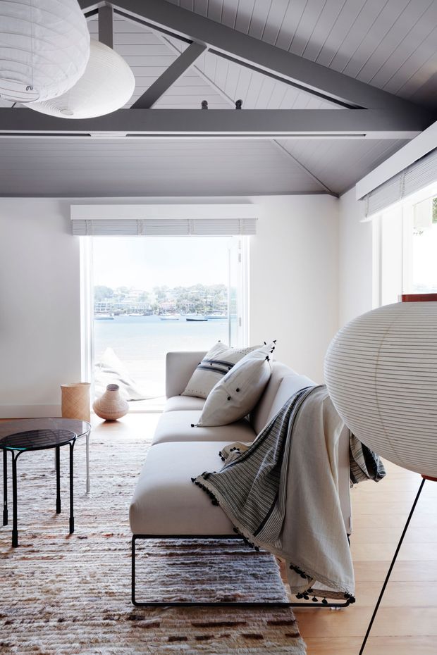 Bright, airy living room with a white sofa, layered neutral cushions and throws, a textured area rug, round glass coffee table, sculptural paper pendant lights, and large windows overlooking a waterfront scene. The room features a dramatic pitched ceiling with exposed dark structural beams and white timber ceiling lining, creating a relaxed contemporary coastal atmosphere.