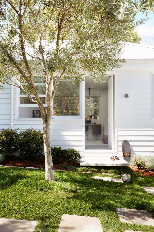 Bright exterior view of a small white weatherboard house with a simple pitched metal roof, large front window, and open entry door. A neat landscaped front garden with lawn, stepping-stone path, shrubs, and a mature tree frames the facade. The interior entry and a compact workspace are visible through the open doorway.