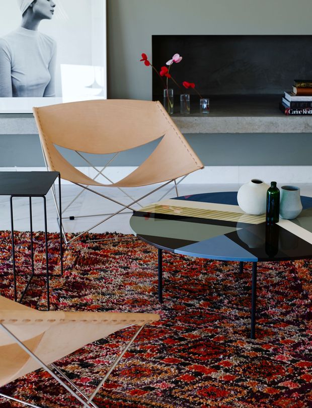A stylish mid-century-inspired living room with two tan leather sling chairs, a low geometric black coffee table, a colourful patterned rug, and sculptural ceramic vases. The room features a dark fireplace or recessed wall opening with a concrete mantel, white built-in cabinetry, minimalist artwork, and a restrained grey-white architectural palette accented by vivid red, orange, blue, and black furnishings.